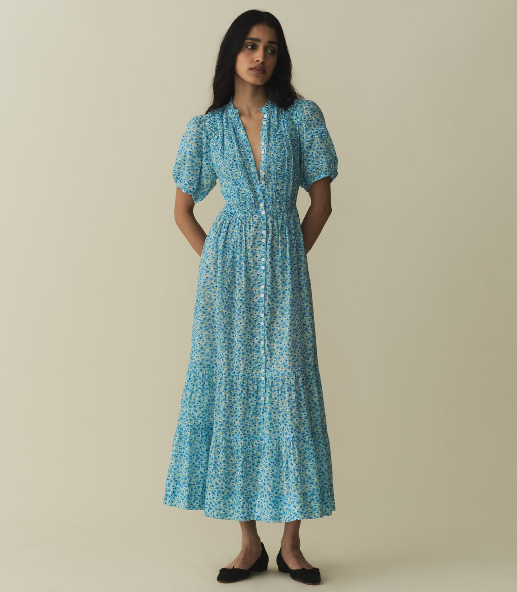 The Haleigh Dress—constructed from 100% organic cotton voile—features a high neck, mother-of-pearl buttons, and short puffed sleeves. Alternating panels of pintucks and ruching lend texture to the fitted bodice, which gives way to a tiered, ankle-grazing A-line skirt graced with clustered pintucks.