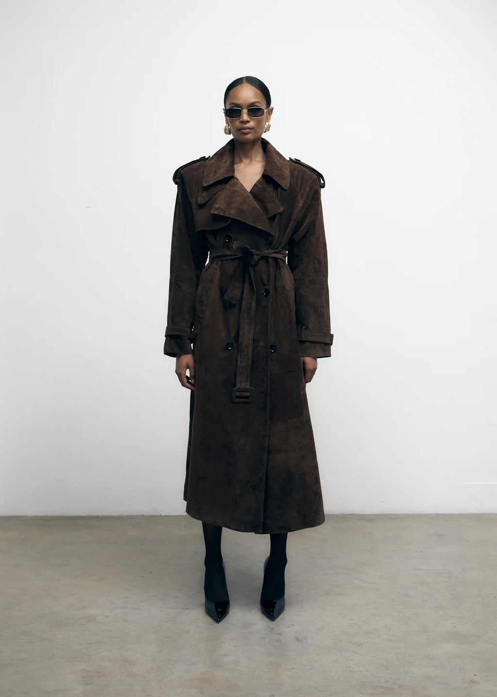 Elevate your wardrobe with the Brown Suede Trench Coat from Desavary London. Crafted with luxurious suede, this double breasted coat boasts an oversized fit and structured shoulders for a look that exudes sophistication. The buckle waist belt and storm flap add practicality to this statement piece. A must-have for any fashion-forward individual.