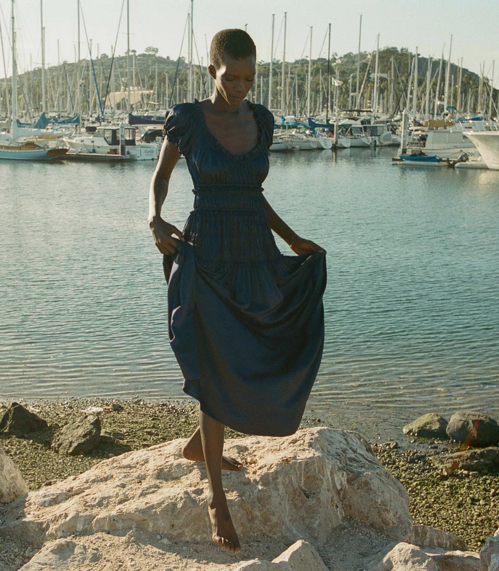 Crafted from a deep navy silk satin, the fan-favorite Leanne Dress features a scoop neckline, a gathered bust, and a fitted bodice framed with petite ruffles. A third gathered panel finishes just below the hips, flaring out into an ankle-length skirt, while elastic lends give to the short puffed sleeves, bust, and waist.