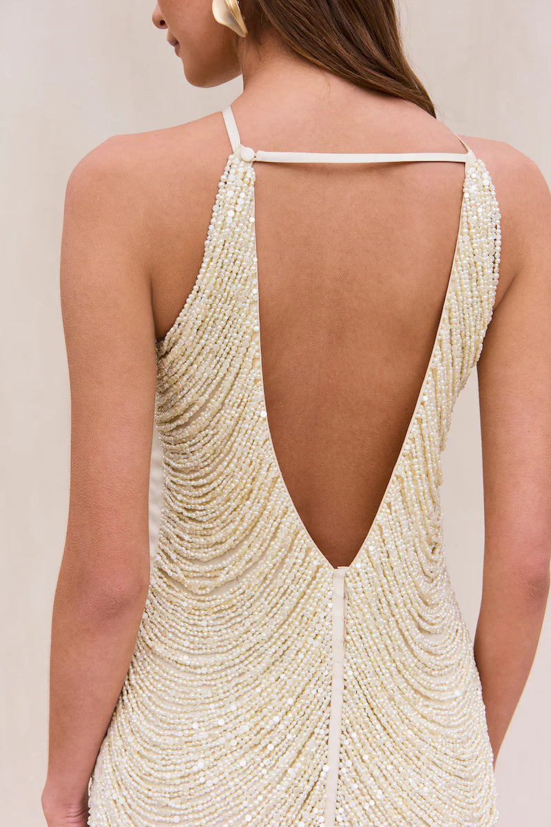 Indulge in the luxurious and exclusive Domina Dress by Cult Gaia. This exquisite halter dress boasts a layered and draped faux pearl fringe that gracefully moves with you. The plunging open back and flattering v-cut silhouette make this mini dress a must-have for any elegant occasion. Elevate your style with this captivating piece.