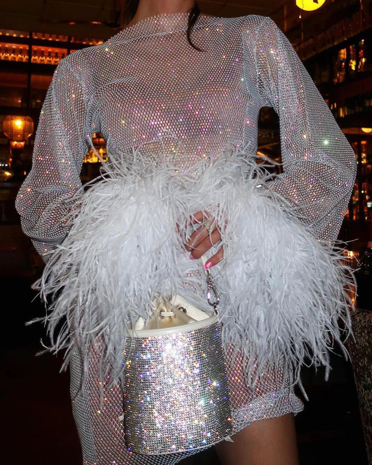 Indulge in the luxurious feel of the Feathers Mini Dress. This mini dress features wide sleeves and an ostrich feather that adds a touch of elegance. Crafted from open knit fabric with rhinestone embellishments throughout, this dress is perfect for making a statement at any event.